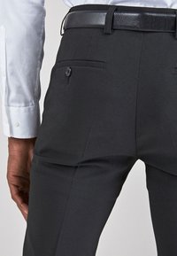 Pantalones negros a medida, elaborados con una tela suave, que cuentan con dos bolsillos traseros con botones y un cinturón de cuero negro que complementa el diseño.