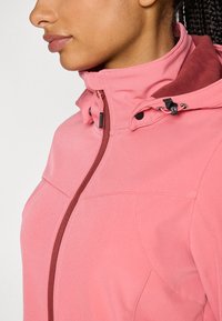 Mujer con chaqueta softshell rosa con cuello alto y capucha ajustable, cremallera parcialmente cerrada, mostrando el perfil desde el cuello hasta el pecho.