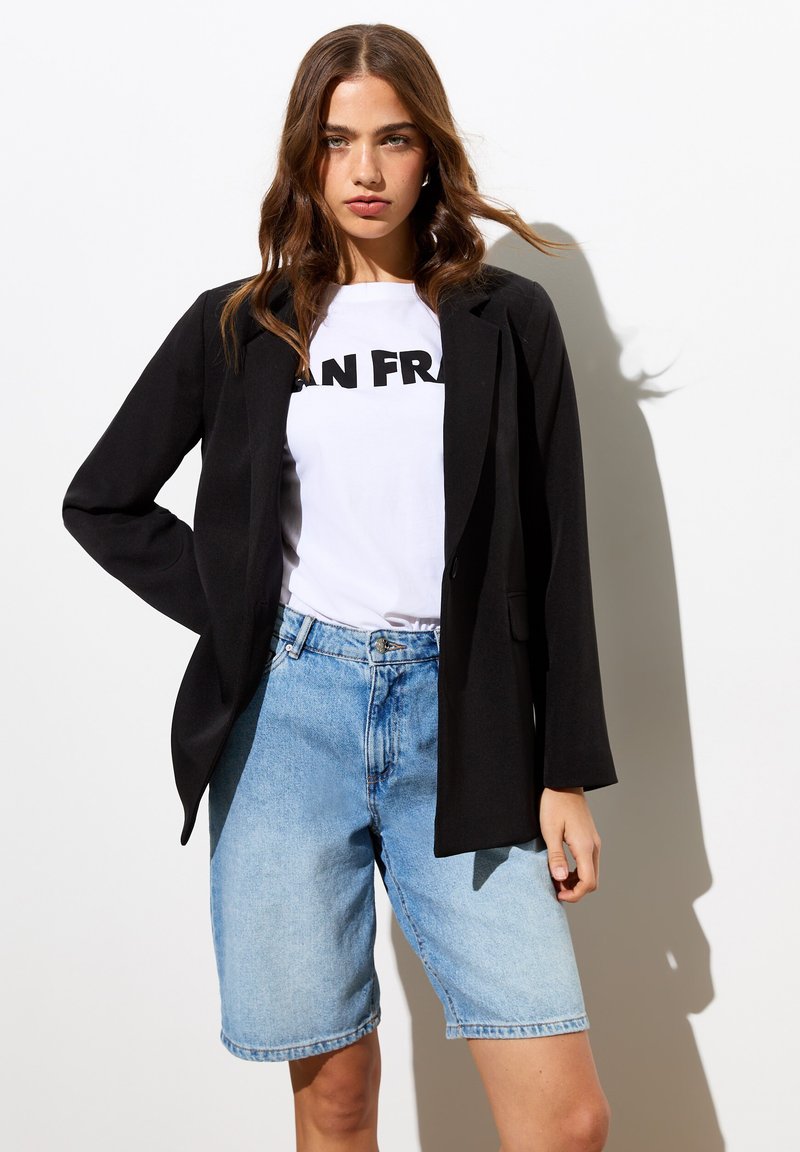 Blazer noir avec une coupe ajustée, associé à un t-shirt blanc portant un texte noir, et un short en denim bleu clair avec une coupe décontractée.
