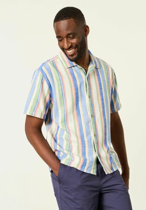 Lächelnder Mann mit den Händen in den Taschen trägt ein kurzärmeliges Hemd mit vertikalen Pastellstreifen und dunkelblaue Hose vor einem schlichten Hintergrund.