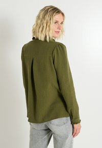 Blusa texturizada verde oliva, de mangas largas, con espalda plisada y puños deshilachados. Presenta un cuello sutil y un corte relajado.
