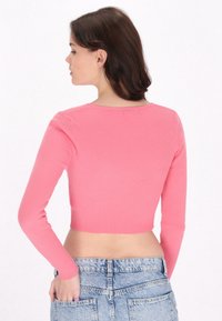 Maglietta a maniche lunghe a coste e corta di colore rosa brillante. Tessuto a maglia testurizzato con scollatura rotonda. Indossata con pantaloni shorts di denim blu chiaro.