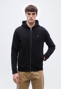 Jeune homme aux cheveux bouclés portant un sweat à capuche noir zippé avec un petit logo et un pantalon beige, debout avec une main dans la poche.