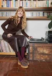 Maglietta marrone a coste con maniche lunghe, gonna in pelle lucida marrone, collant bordeaux, scarpe piattaforma grigie con lacci rossi, cassa di legno, dischi in vinile.