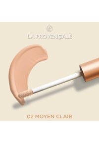 Correcteur liquide en teinte beige clair, avec une texture douce et crémeuse sur un swatch courbé. L'emballage présente un capuchon en cuivre mat et un applicateur blanc.