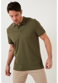 Polo de color verde oliva con una tela texturizada de canalé, tapeta de tres botones y mangas cortas. Presenta un sutil detalle de costura en el collar.
