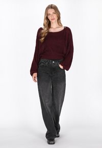 Burgundowa dziana sweter z długimi, luźnymi rękawami, w parze z czarnymi spodniami o wysokim stanie; model nosi czarne buty, stoi z rękami w kieszeniach.
