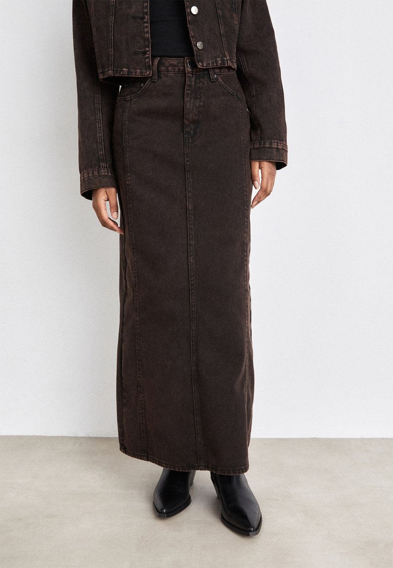 Femme portant une jupe maxi en denim marron foncé, des bottes noires et une veste courte en denim assortie sur un fond clair uni.
