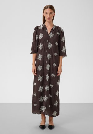 Femme portant une longue robe marron foncé avec des motifs floraux blancs, des manches trois-quarts et des chaussures pointues noires sur un fond uni.