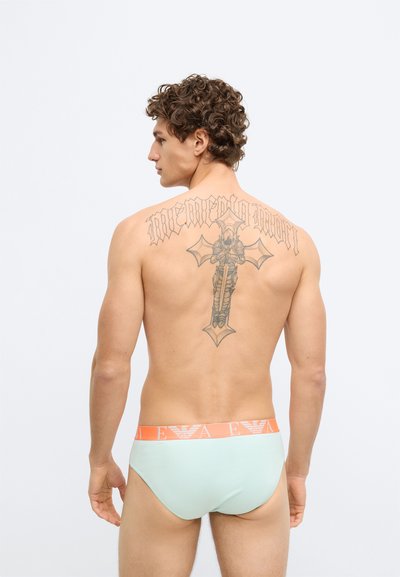 Cuecas verdes menta com cintura alaranjada e logotipo, apresentando uma textura suave. Design de tatuagem notável visível na parte superior das costas.