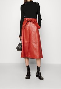 Czerwona skórzana spódnica midi z przednim paskiem wiązanym na kokardę, zestawiona z czarną bluzką z długim rękawem i czarnymi lakierowanymi botkami za kostkę. Elementy podkreślają czyste linie.