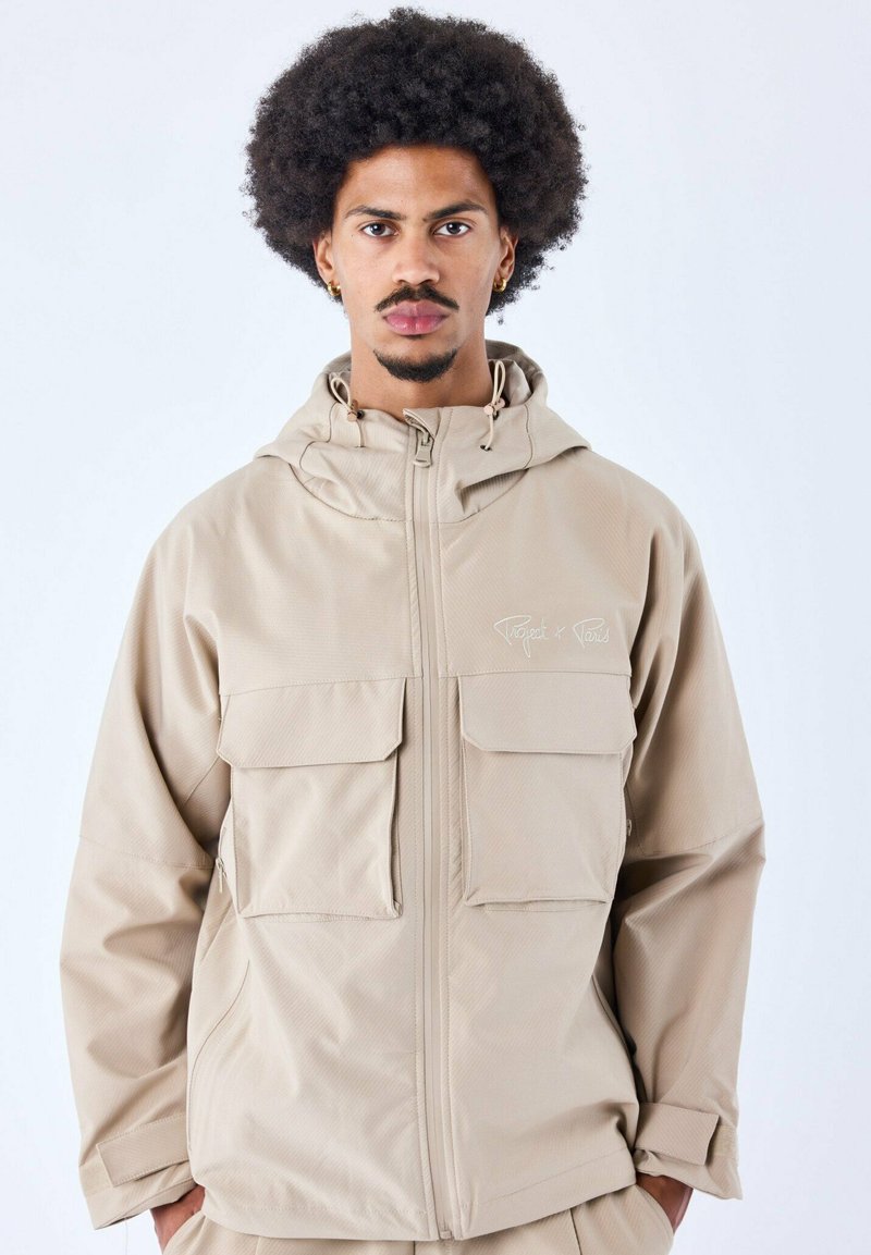 Chaqueta beige con cremallera y capucha. Presenta dos amplios bolsillos frontales y una textura sutil. Material impermeable, diseñado para uso al aire libre.