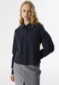Camicia blu navy con bottoni e colletto, due tasche sul petto e maniche a palloncino. Il tessuto appare liscio e leggero, con una vestibilità comoda.