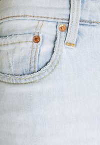 Tissu en denim bleu clair avec une poche visible, des rivets en cuivre et des détails de couture jaune le long des coutures et des bords.