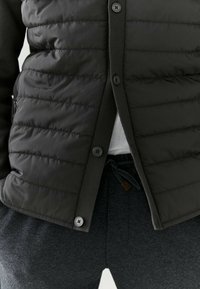 Padded dark gray jacket with a buttoned front, featuring a smooth texture and ribbed knit cuffs. Worn over gray drawstring pants.