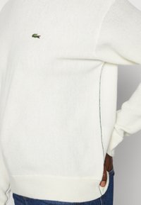 White knitted sweater with ribbed hem and cuffs. Features green accent stitching and a small embroidered logo on the back.