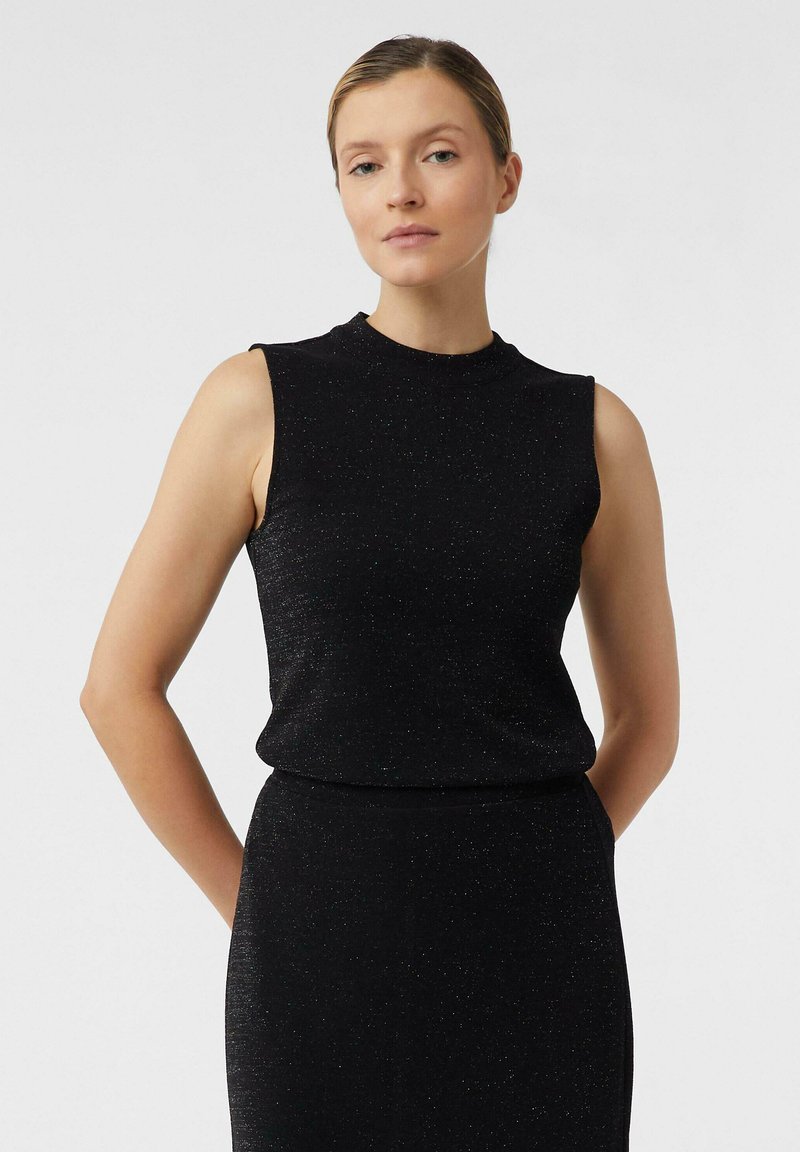 Eine Frau steht mit den Händen hinter dem Rücken in einem ärmellosen, taillierten schwarzen Kleid mit dezentem Schimmer vor einem schlichten hellen Hintergrund.