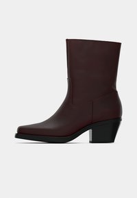 Botín de cuero marrón oscuro con un tacón bajo y de bloque, y punta afilada, mostrado en perfil lateral sobre un fondo neutro.