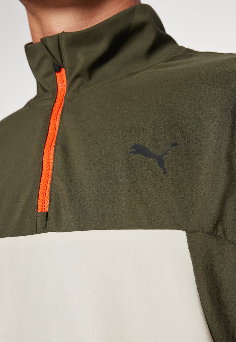 Olijf groene half-zip jack met een oranje rits, voorzien van een zwart Puma-logo op de borst en een crèmekleurig paneel aan de onderkant. Zachte textuur.