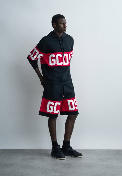 Άνδρας στέκεται μπροστά σε απλό τοίχο φορώντας μαύρο και κόκκινο φούτερ με κουκούλα και ταιριαστά σορτς με λευκά γράμματα "GCDS", μαύρες κάλτσες και αθλητικά παπούτσια.