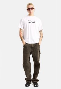 Hvid t-shirt med korte ærmer og sort "VANS"-logo, kombineret med mørkebrune baggy jeans og sorte sneakers. Modellen bærer også mørke solbriller.