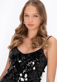 Fekete flitteres ruha, mélyen kivágott nyakkivágással és vékony pántokkal, csillogó, kör alakú flitterekkel díszítve, amelyek visszaverő textúrát kölcsönöznek neki.