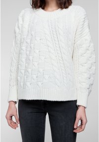 Vit kabelstickad tröja med rund halsringning, texturerat mönster och ribbade ärmmuddar. Buren med mörka jeans, vilket betonar kontrasten.