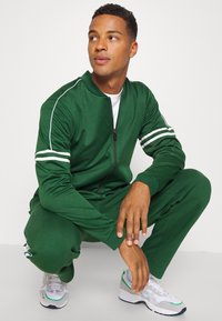 Grüner Trainingsanzug mit weißen Akzenten, Reißverschlussjacke und schmal zulaufenden Hosen, aus weichem Stoff. Modell in grauen Sportschuhen in der Hocke.