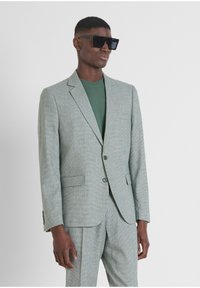 Traje texturizado gris claro con solapa de muesca, dos botones y bolsillos laterales, combinado con una camiseta verde de cuello redondo y gafas de sol negras.