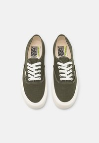 Zapatillas de lona verdes con cordones blancos, suelas de goma y interior beige. Presentan bordes cosidos y una etiqueta de logo en la lengüeta.