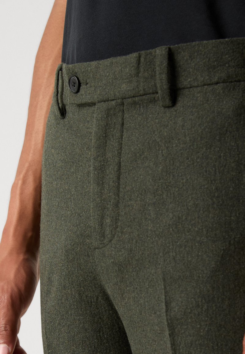 Dunkelgrüne Wollhose mit glatter Textur, versehen mit einem Knopfverschluss, seitlichen Taschen und einem taillierten Design. Sichtbare Nähte setzen Akzente.