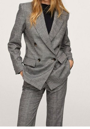 Woman wearing gray plaid double-breasted blazer and matching pants with hands in pockets, black top underneath, standing against neutral background.