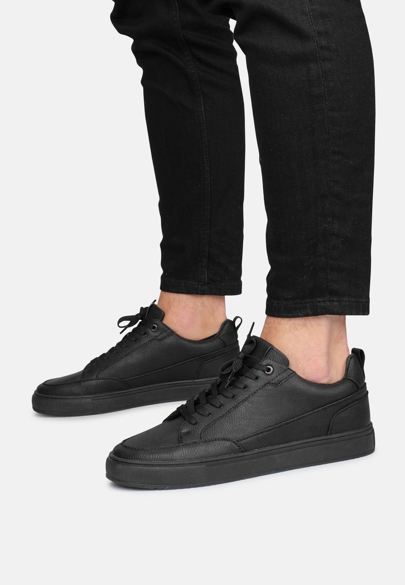 Baskets basses en cuir noir avec lacets, portées avec un pantalon noir remonté, dévoilant les chevilles sur un fond clair uni.