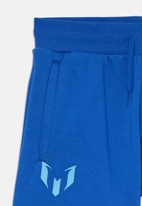 Mėlyni sportiniai kelnės, pagamintos iš minkštos medžiagos, su elastingu juosmeniu, šoniniais kišenėmis ir šviesiai mėlynu logotipo akcentu ant kojos.