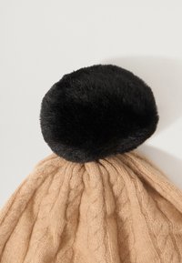 Knit beige beanie with a textured cable pattern, featuring a black faux fur pom-pom at the top. Soft material with a relaxed fit.