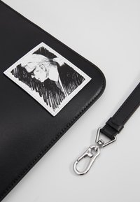 Černá kožená taška s bílou nášivkou obsahující černou ilustraci. Obsahuje stříbrný kovový klip pro připevnění. Elegantní a hladký design.