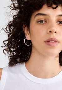 Pendiente de aro de plata con un acabado suave y brillante. Primer plano de un modelo con cabello rizado, que lleva una blusa sin mangas blanca.