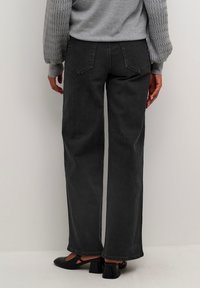 Jean large noir en denim, avec un design classique à cinq poches et des détails de couture subtils. Porté avec un pull gris et des chaussures noires.
