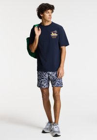 Camiseta azul marino con un diseño coral, combinada con pantalones cortos de patrón en azul marino y blanco. Incluye una bolsa de mano verde y zapatillas grises.