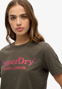Camiseta verde oliva de manga corta con un gran logo brillante en rosa y púrpura de "SuperDry" y el texto "SOHO.LONDON." debajo.