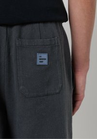 Detailný záber na tmavosivé vrecko nohavíc s modrou nálepkou "LA MAISON AMV", ktoré sú kombinované s čiernou košeľou, pričom je vidieť pravú ruku osoby.