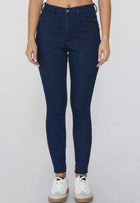 Jeans ajustados de color azul oscuro hechos de denim, con un clásico diseño de cinco bolsillos y cierre de botón en la parte delantera. Combinados con zapatillas blancas.