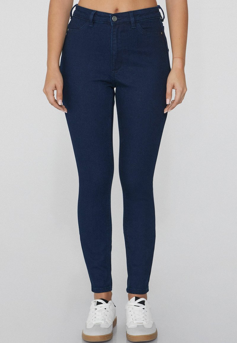 Jeans ajustados de color azul oscuro hechos de denim, con un clásico diseño de cinco bolsillos y cierre de botón en la parte delantera. Combinados con zapatillas blancas.