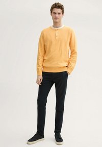 Camiseta henley amarilla de manga larga con tres botones y puños acanalados, combinada con pantalones oscuros y zapatillas negras. Tejido suave y ligero.