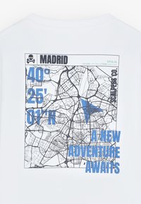 Witte t-shirt met een grafische kaart van Madrid, met blauwe tekst die coördinaten weergeeft en de zin "EEN NIEUWE AVONTUUR WACHT."