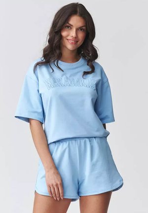 Kobieta uśmiechnięta, ubrana w jasnoniebieską, luźną koszulkę z napisem "MAKADAMIA" oraz dopasowane szorty na jednolitym jasnoszarym tle.