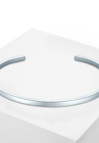 Collar de metal plateado con un diseño liso y redondeado. Presenta una forma elegante y minimalista, con un acabado brillante, reposando sobre una superficie blanca.