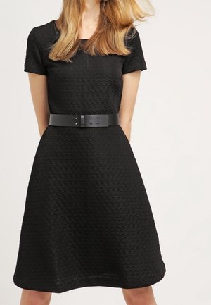 Vrouw draagt een zwarte, textuurrijke jurk met korte mouwen en een brede zwarte riem, staand tegen een eenvoudige lichte achtergrond.