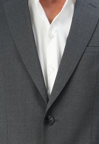Giacca da uomo grigia con rever a punta, un singolo bottone nero e una leggera trama, abbinata a una camicia bianca con bottoni ben stirata.