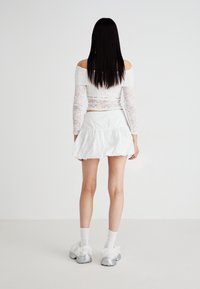 Top bianco in pizzo a maniche lunghe, design off-shoulder, abbinato a una mini gonna bianca a balze. Sneakers argento con dettagli bianchi soffici.